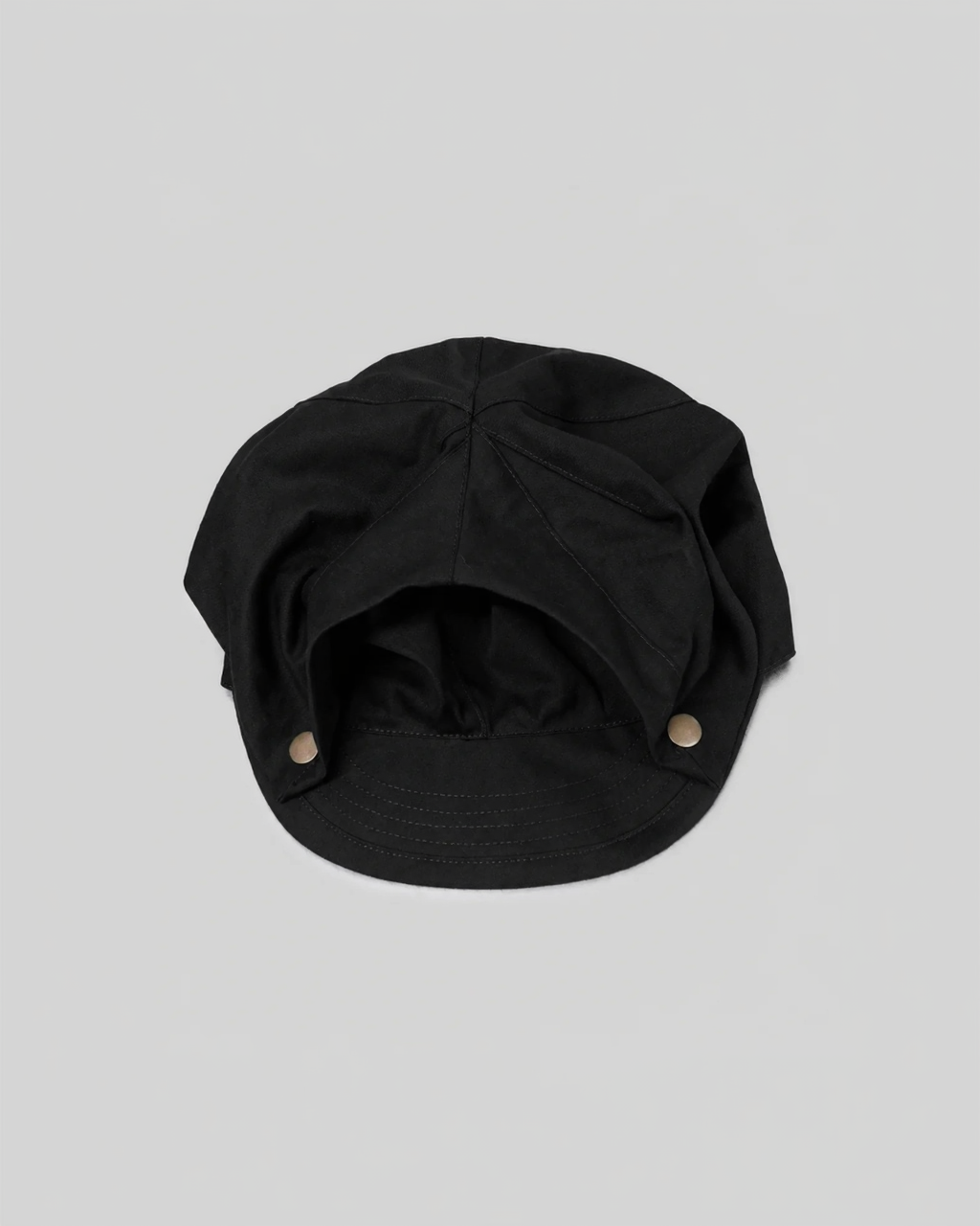 A black fabric cap with a curved brim and two small metallic snap buttons on the sides, photographed against a light gray background.