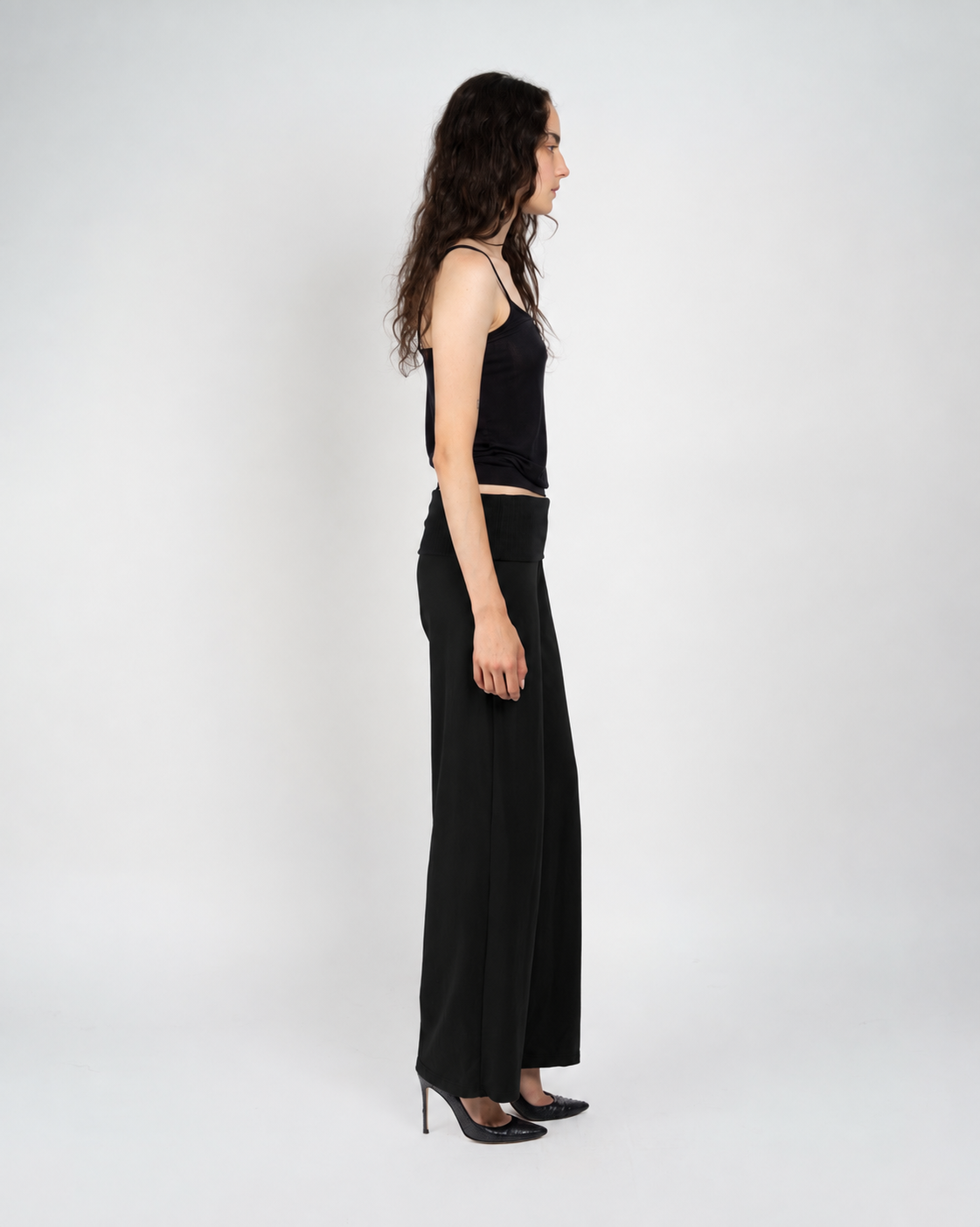 A woman stands in profile wearing a black sleeveless top and black wide-leg pants against a plain light gray studio background.