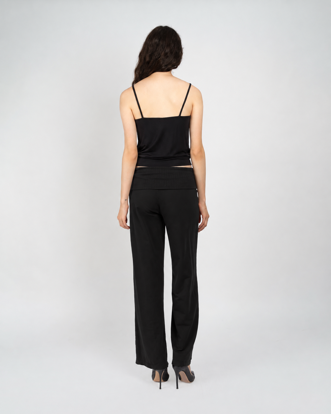 A woman viewed from the back wearing a black camisole top with thin straps and matching black wide-leg pants, standing against a light gray studio background.