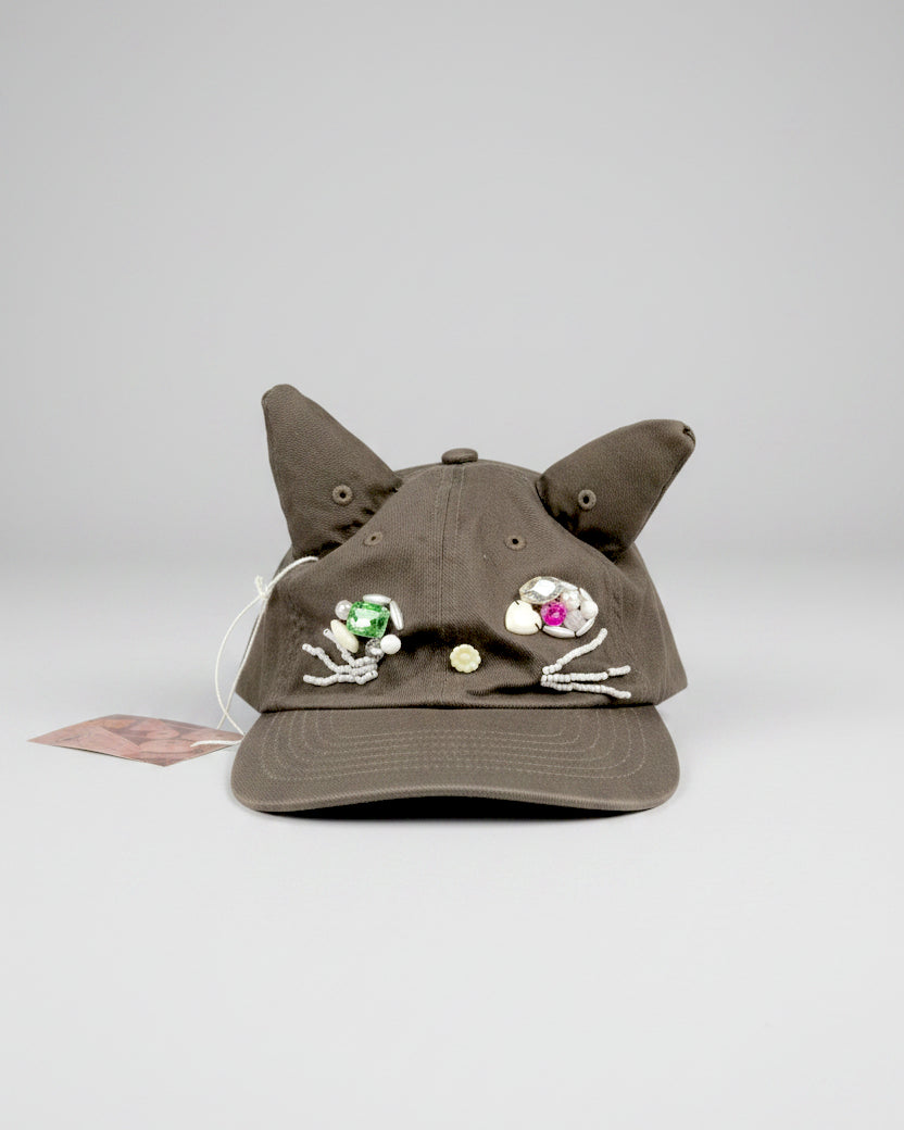 Brown baseball cap with sculptural cat ears and beaded embellishments forming a face, shown against a light gray background.