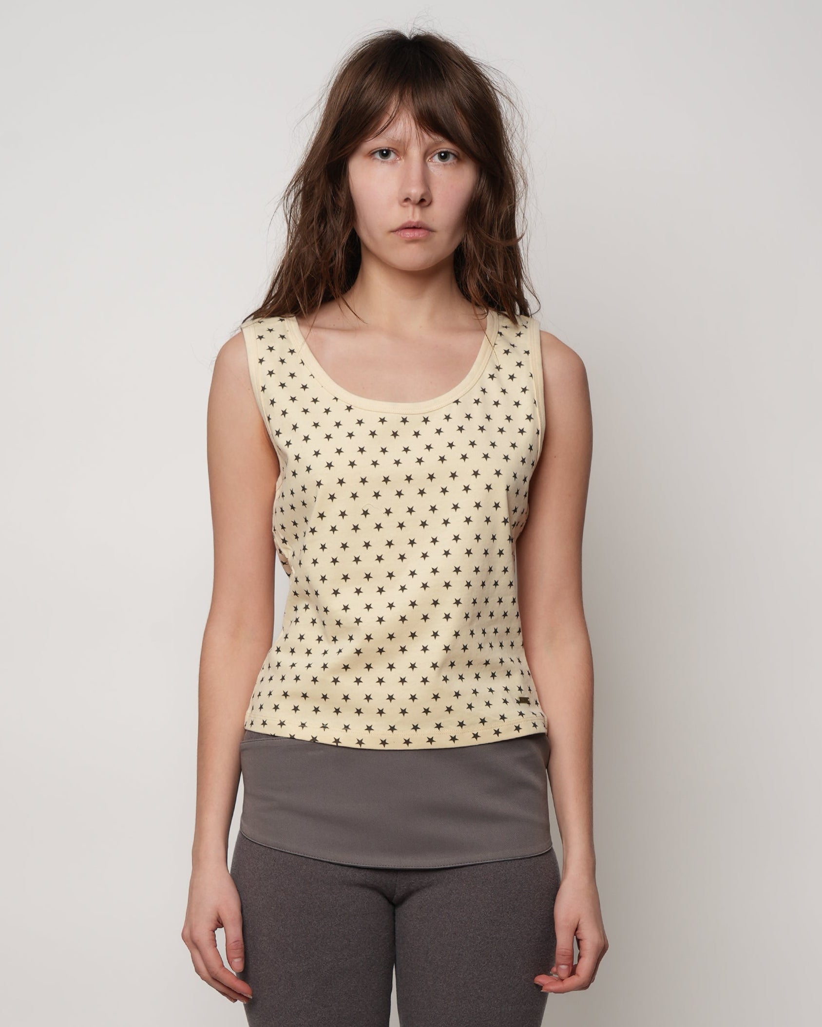 Woman wearing a cream tank top with black polka stars and gray pants on a plain background