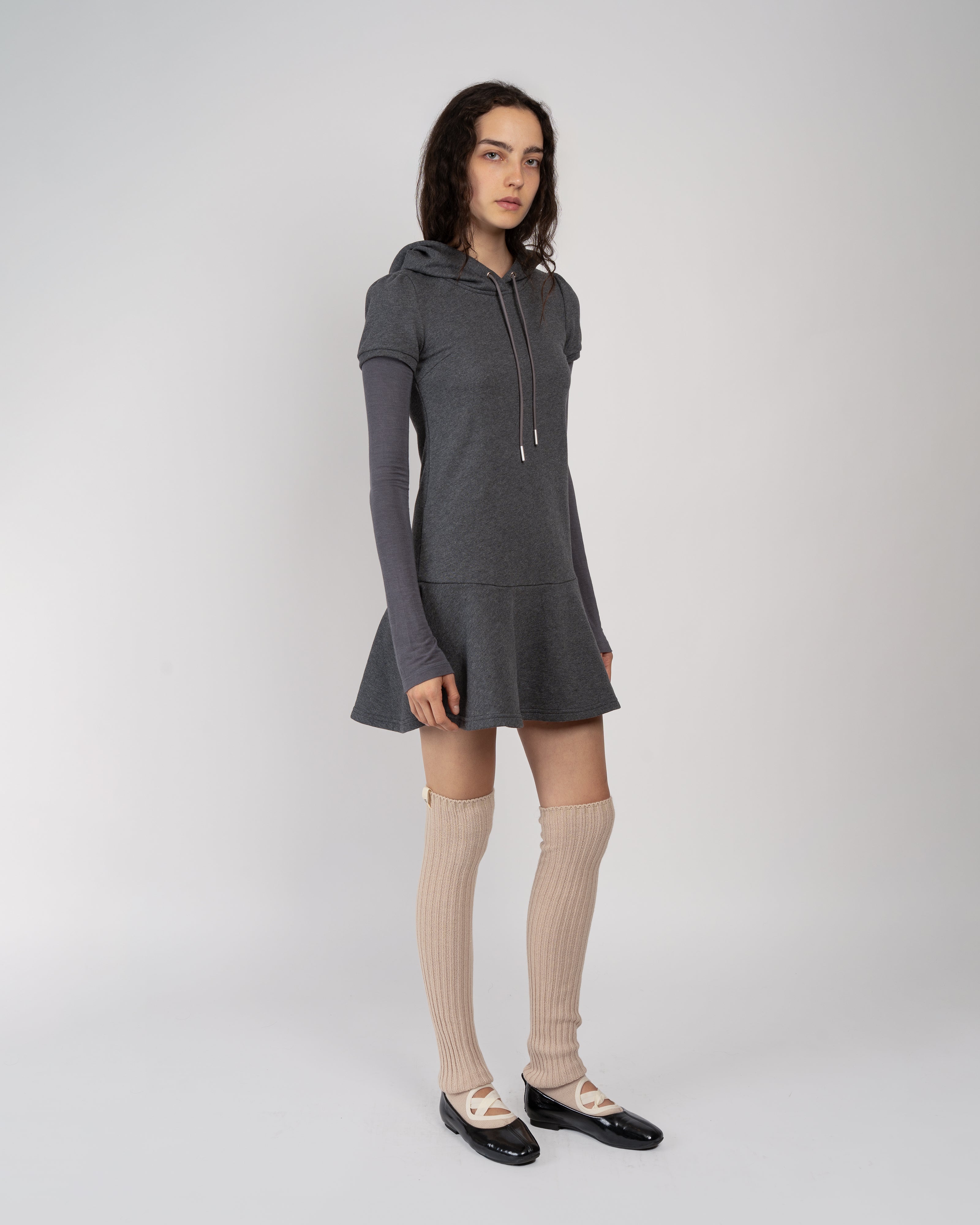 A model stands against a light gray studio background wearing a dark gray hooded long-sleeve mini dress, beige ribbed knee-high socks, and black Mary Jane shoes.