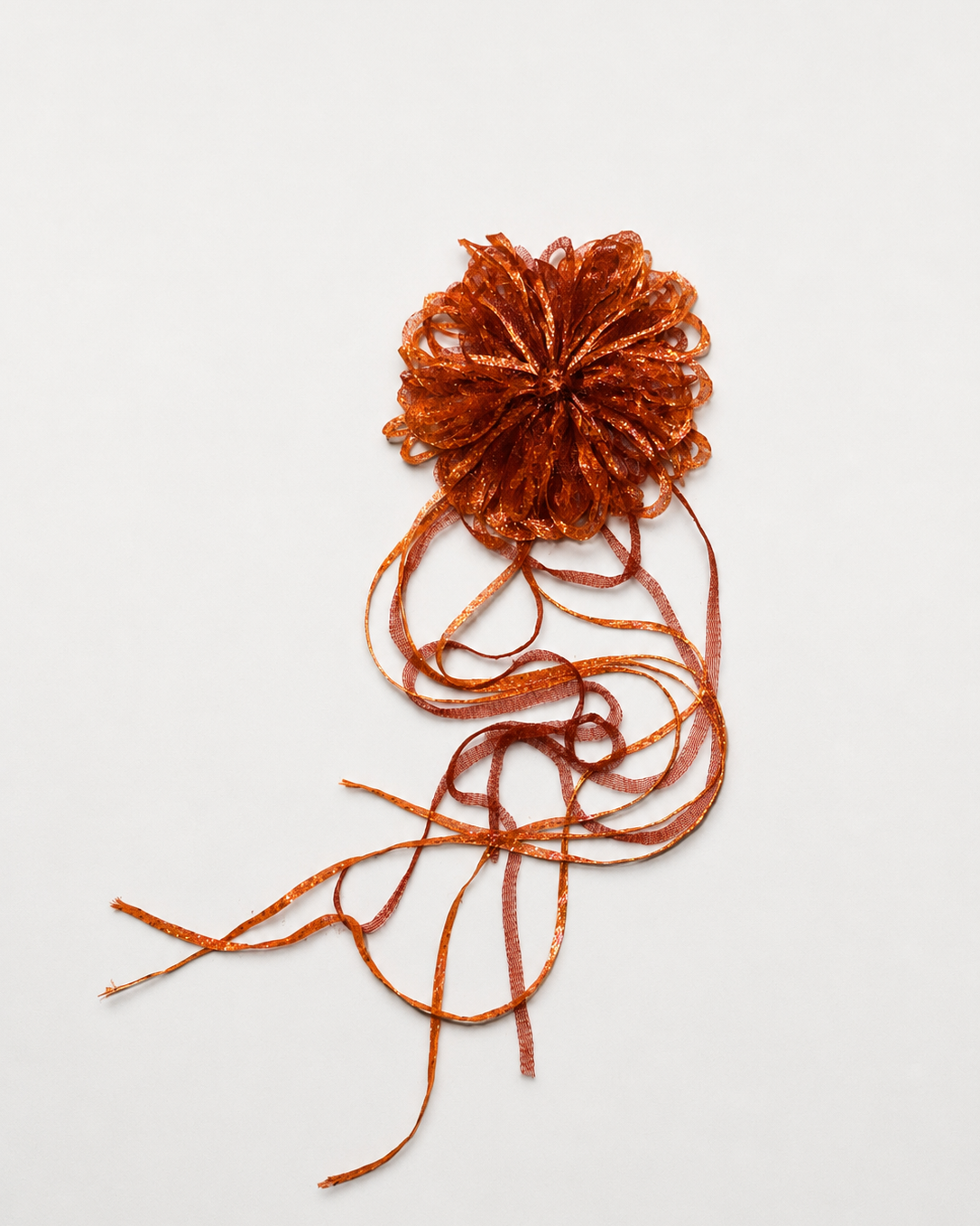 An orange glittery pom-pom hair accessory with long, flowing ribbon-like strands, shown on a plain light gray background.