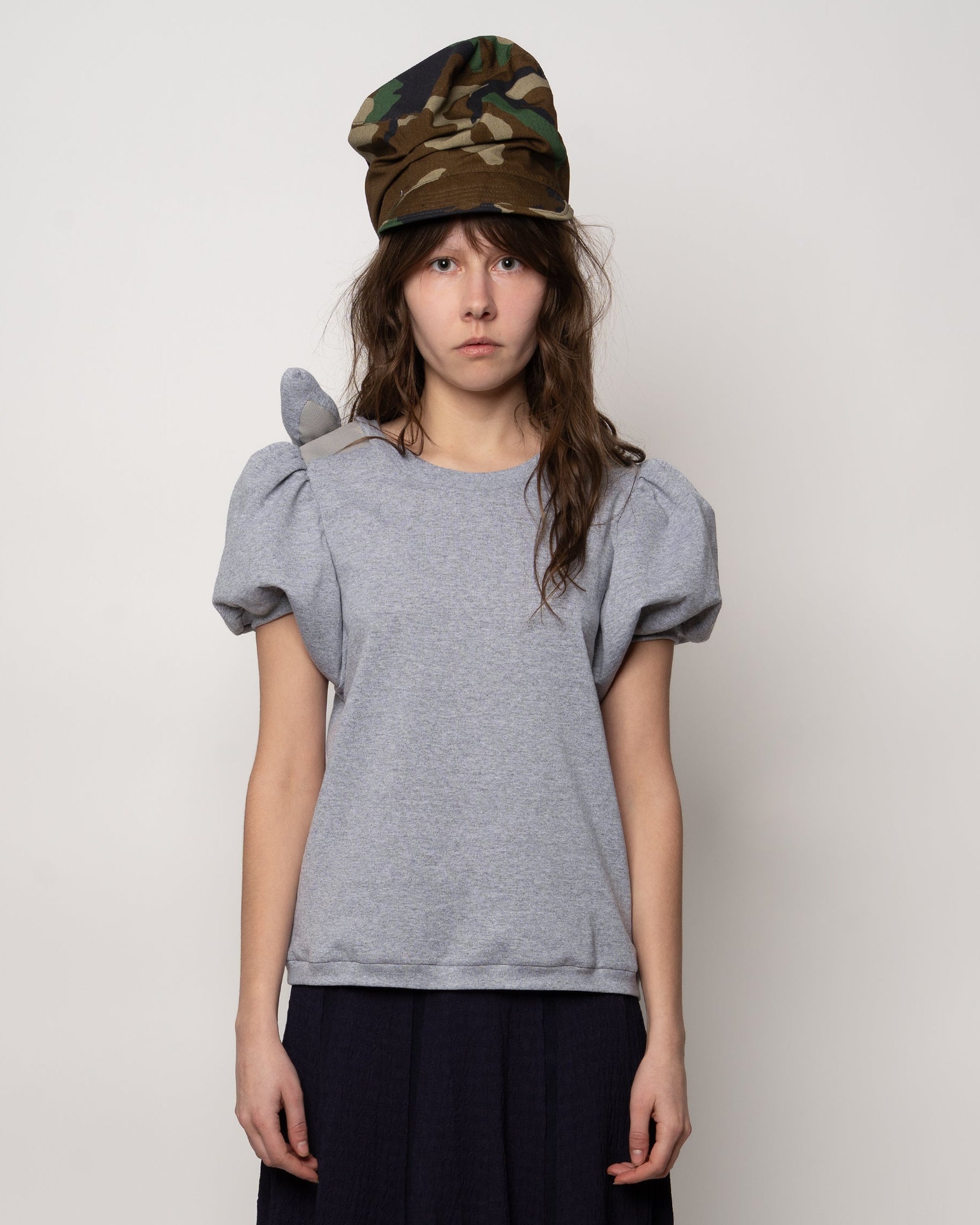 Person wearing a gray t-shirt with puff sleeves and a camouflage hat on a plain background