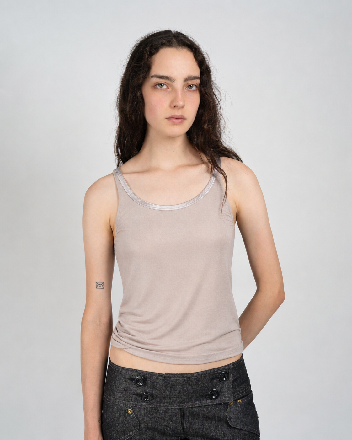 A woman modeling a light beige ribbed tank top with a contrasting gray trim along the scoop neck, paired with dark gray jeans, against a plain light gray studio background.