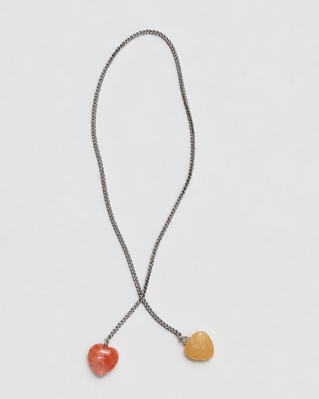 A silver-toned chain necklace with two dangling heart-shaped pendants, one in orange-red and one in pale yellow, displayed on a light gray background.