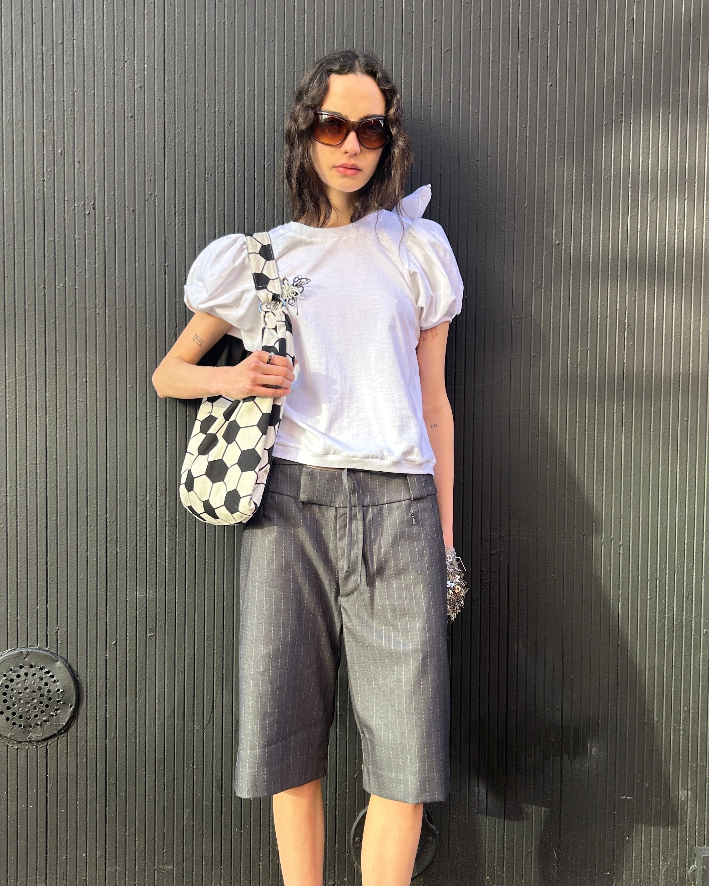 Woman in a white top and gray shorts holding a polka dot bag against a dark wall.